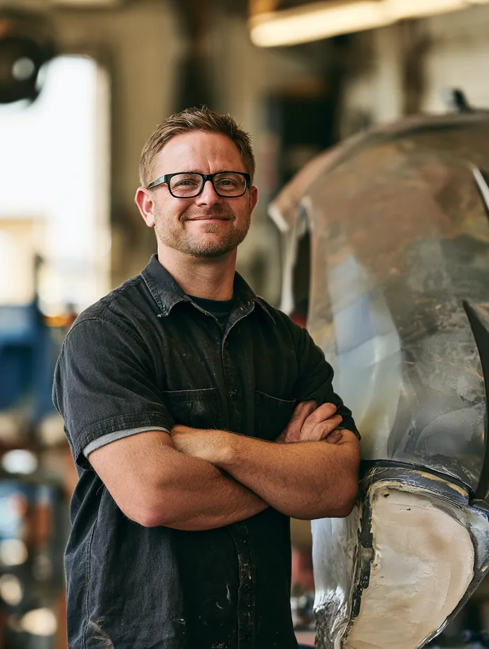 American body shop owner standing beside a half-sanded car fender with arms crossed